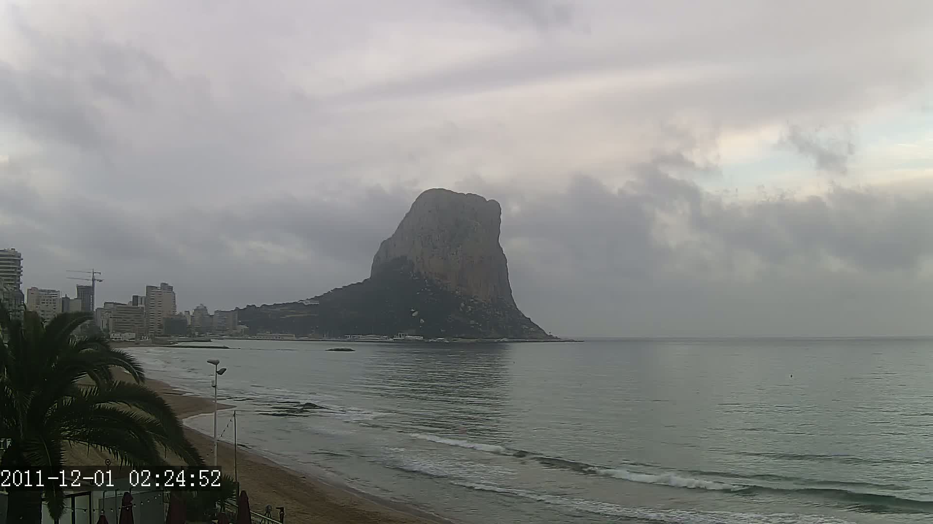 A large, solitary rock formation stands prominently in the distance across a calm, gray sea under an overcast sky, with a sandy beach and a city skyline visible in the foreground.