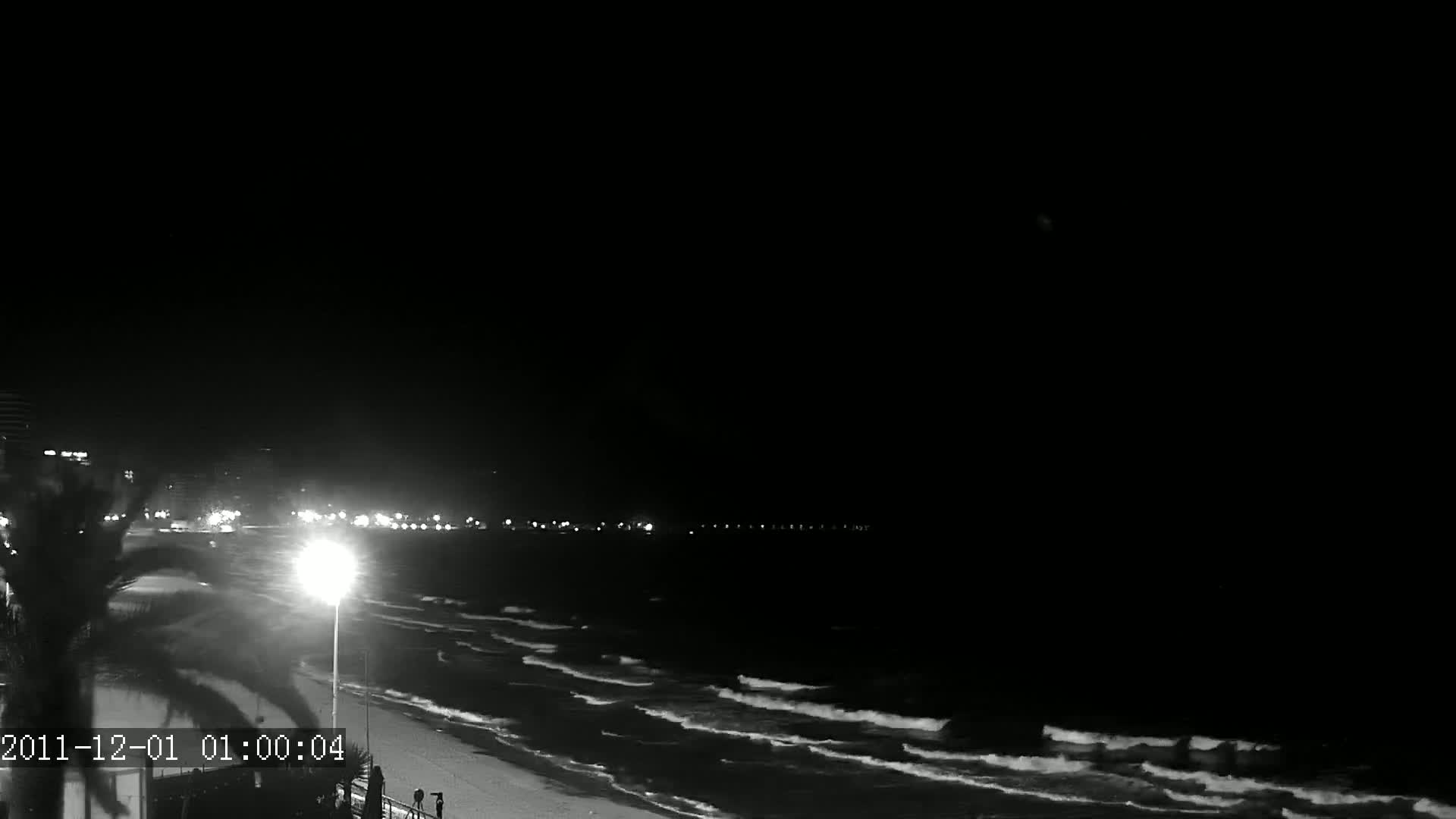 A nighttime beach scene shows gently rolling waves lapping a sandy shore under a dark, clear sky, with lights visible in the distance along a beachfront road.
