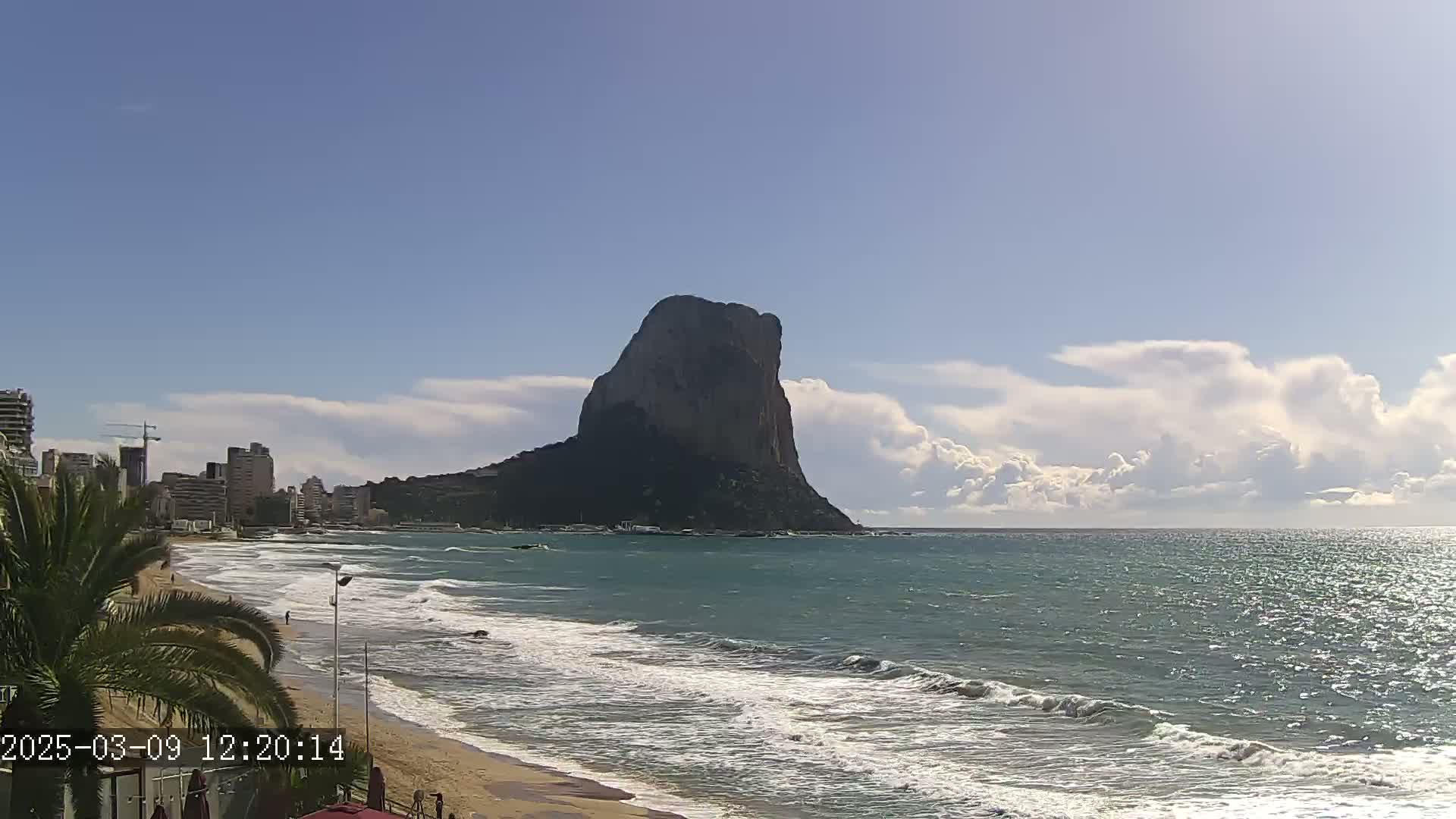 A large, dark rock formation rises from the sea under a partly cloudy sky, with waves lapping a sandy beach in the foreground and buildings visible along the shore.