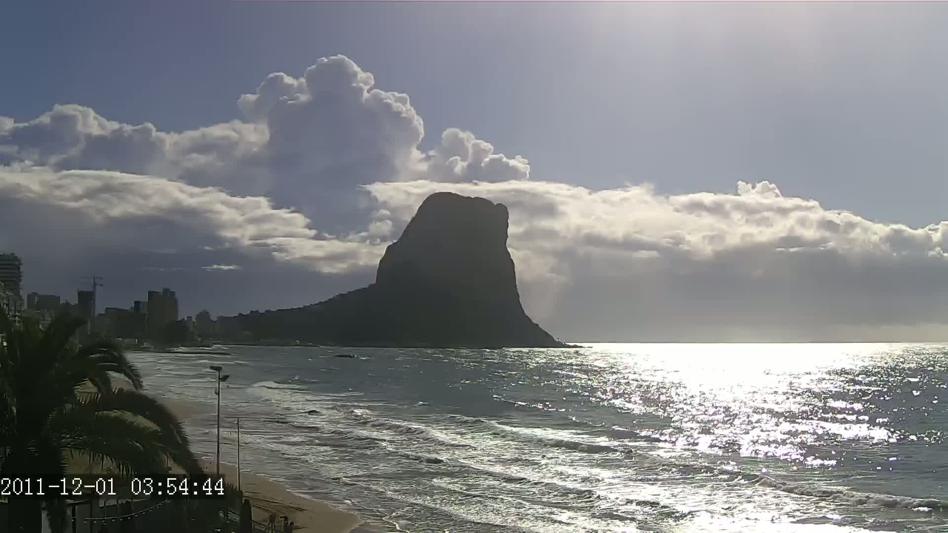 A large, dark mountain stands prominently against a partly cloudy sky over a beach with gentle waves and a city skyline in the distance.