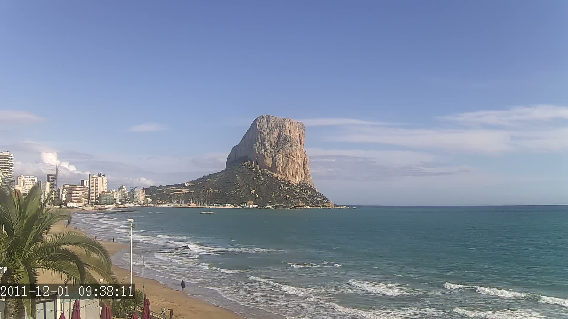 A sandy beach with gentle waves laps the shore, overlooking a calm sea with a large, imposing rock formation in the distance under a mostly clear sky.