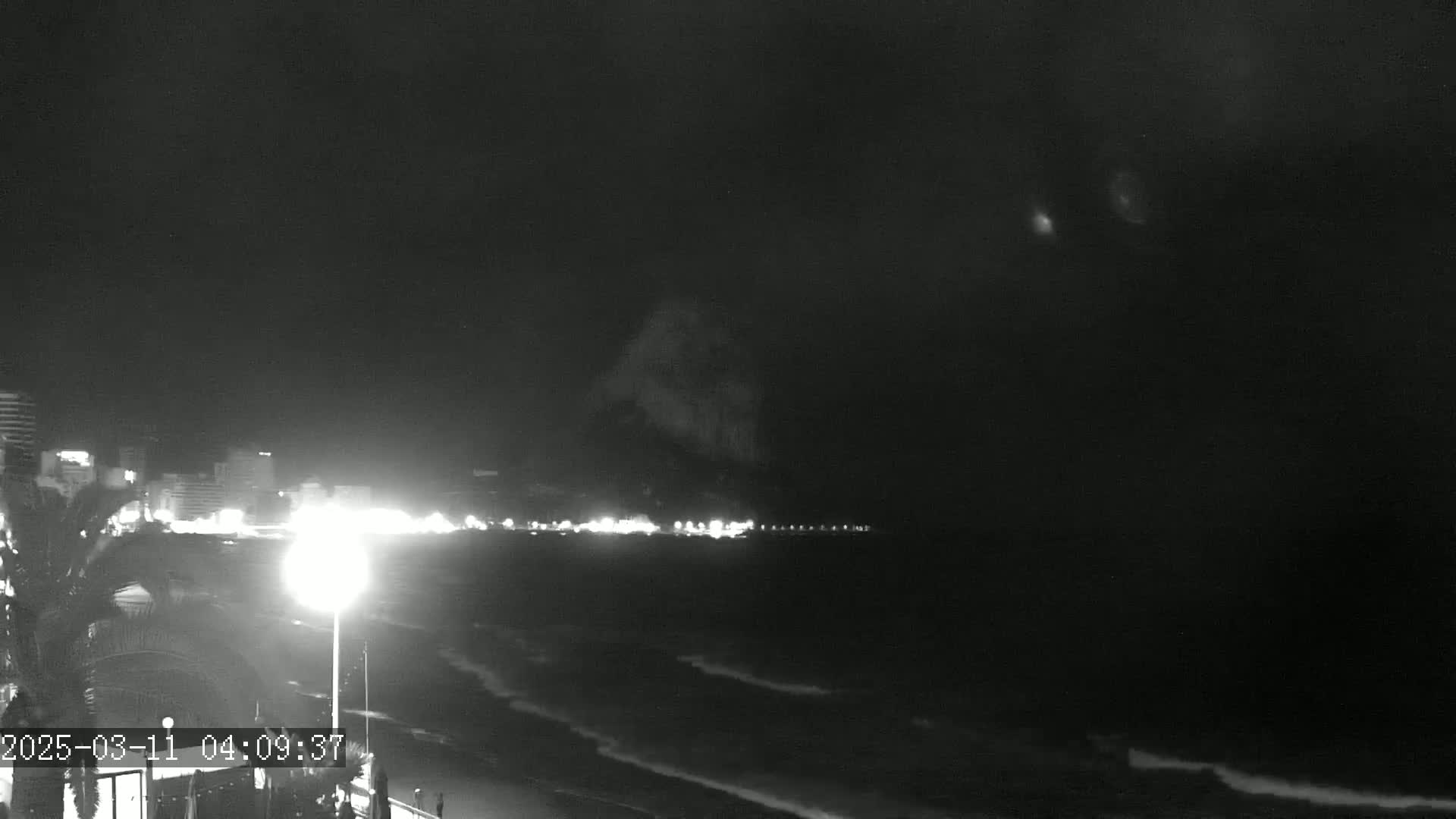 A nighttime view of a dimly lit beach with a city skyline in the distance and gently lapping waves.