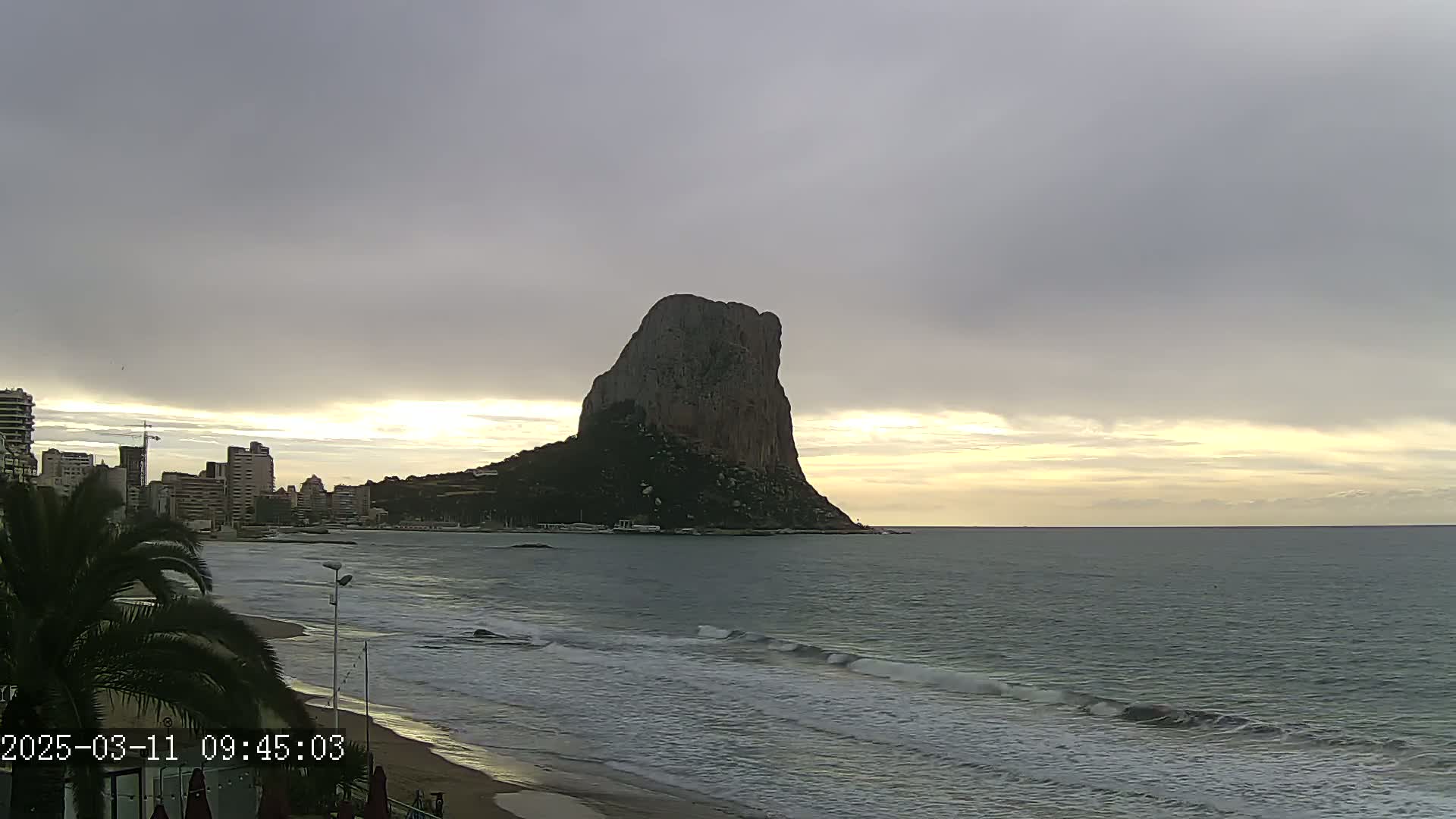 A large, dark rock formation rises from the sea under a cloudy, overcast sky, with a beach and cityscape visible in the foreground.