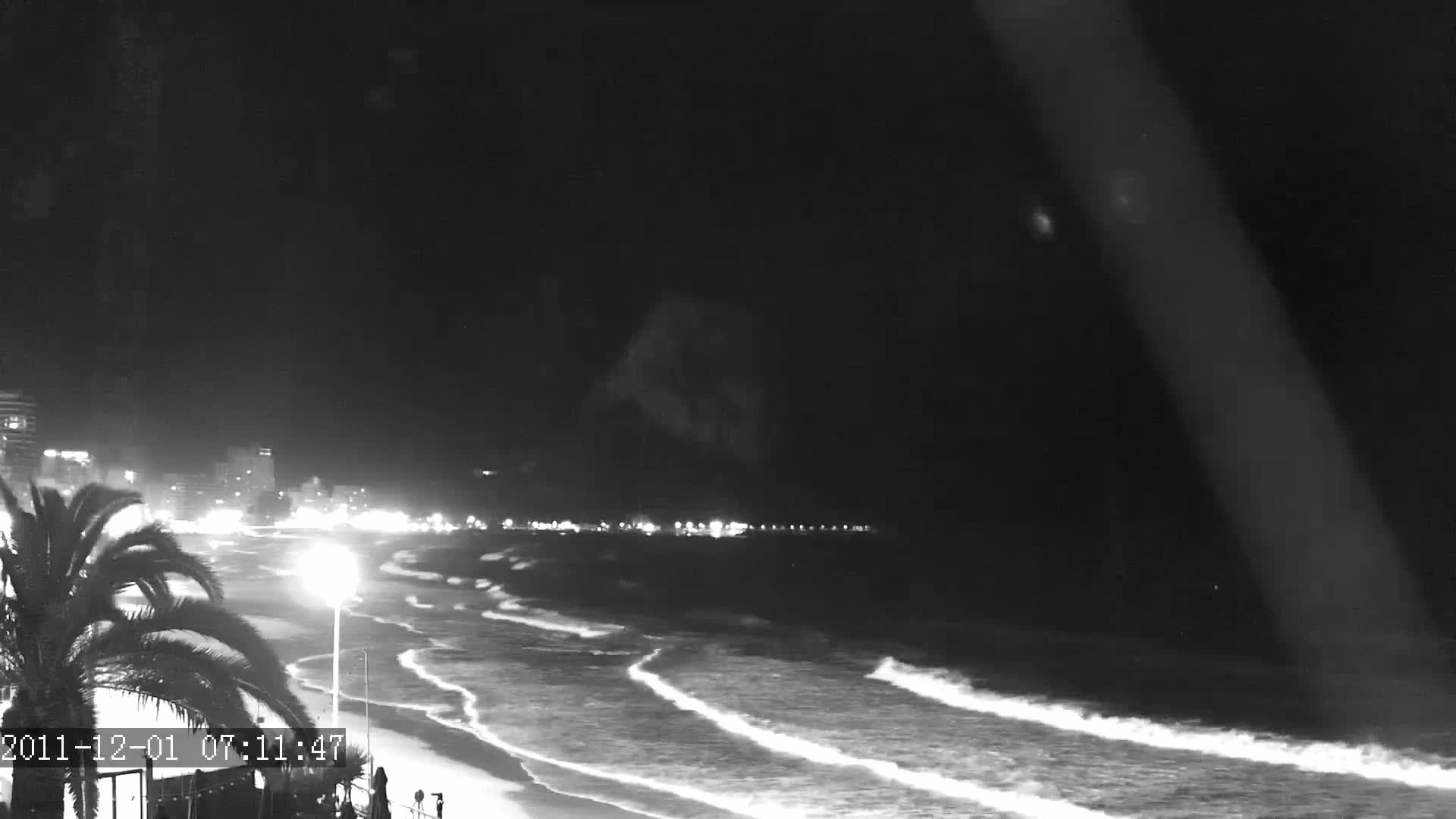 A nighttime, black and white view of a beach with gently rolling waves, illuminated by city lights in the distance under a clear, dark sky.