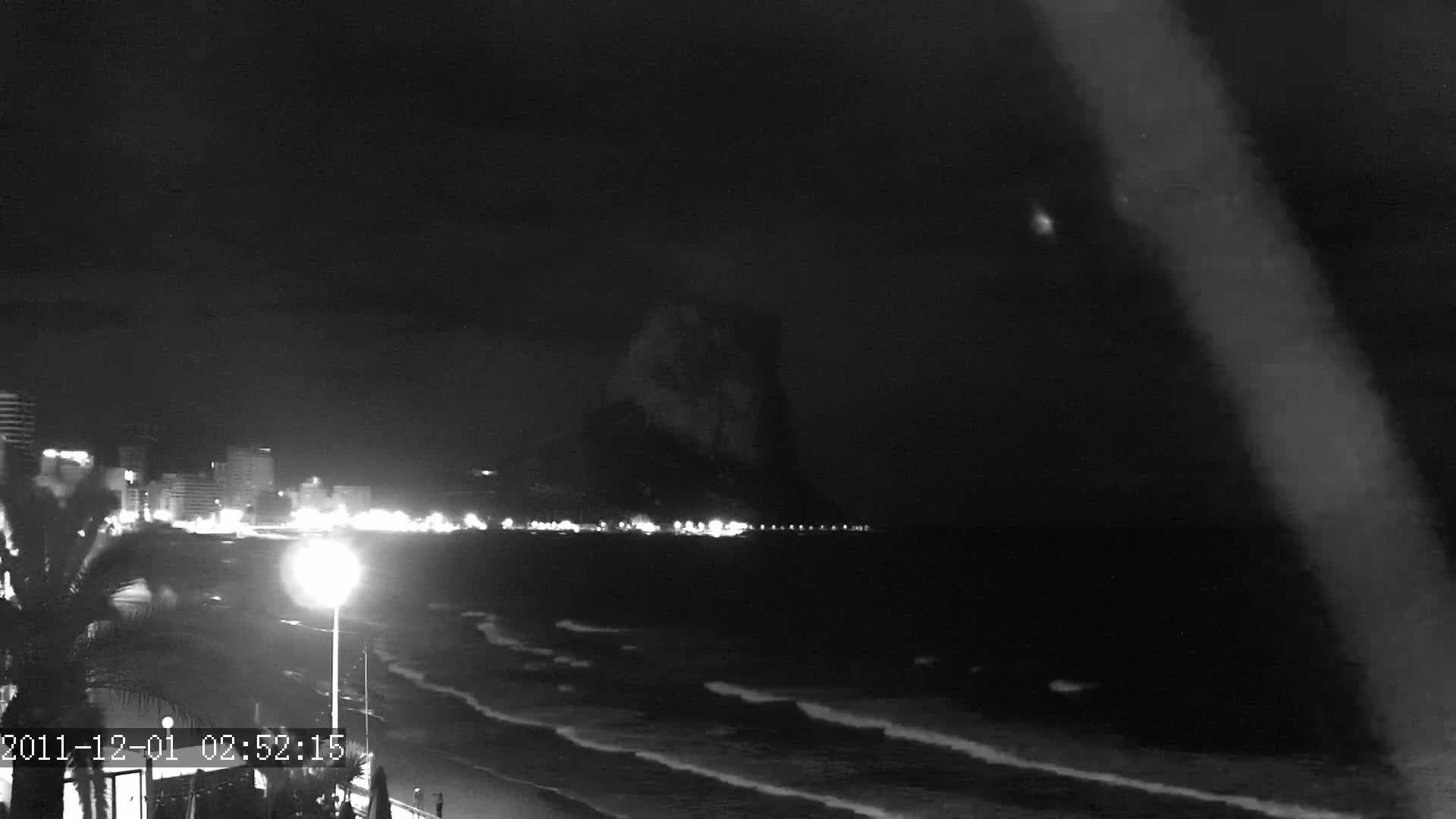 A nighttime beach scene shows a dark, rocky headland looming over a gently waving ocean, with a line of lights from buildings visible along the shore.