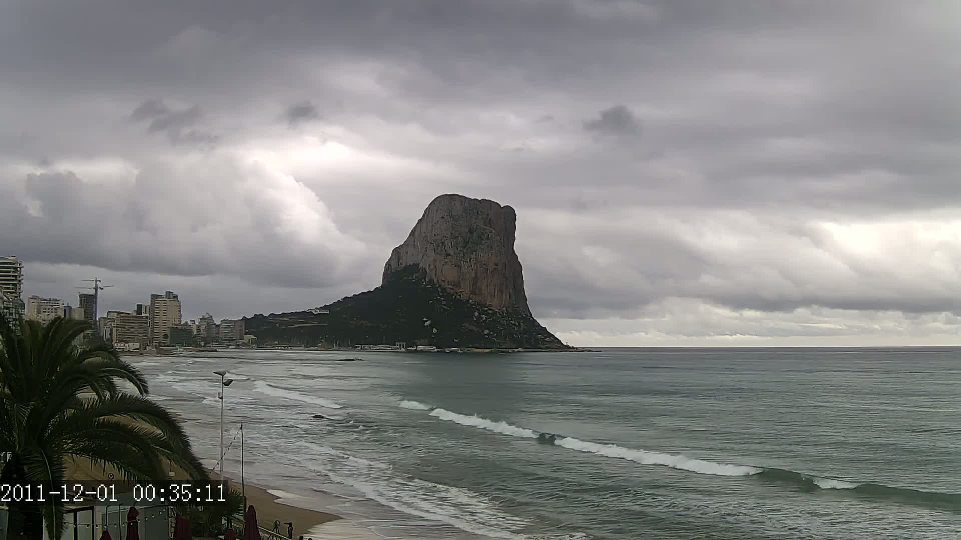 A large, rocky cliff stands prominently offshore, viewed from a sandy beach under a gray, cloudy sky with gentle waves rolling onto the shore.