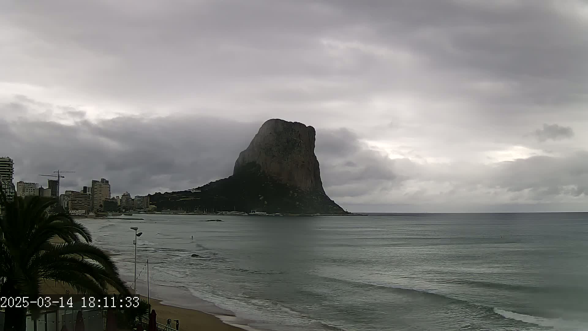 A large, dark rock formation rises from the sea under an overcast sky, with a city skyline and beach visible in the foreground.