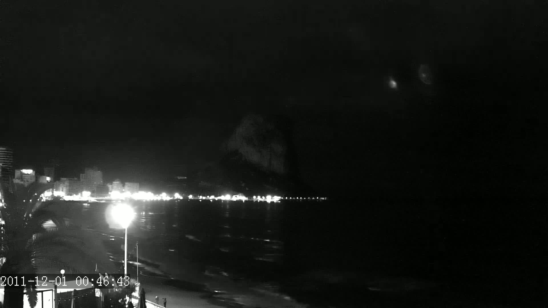A nighttime coastal scene shows a dark body of water with a dimly lit beachfront and buildings in the background under a clear, dark sky.