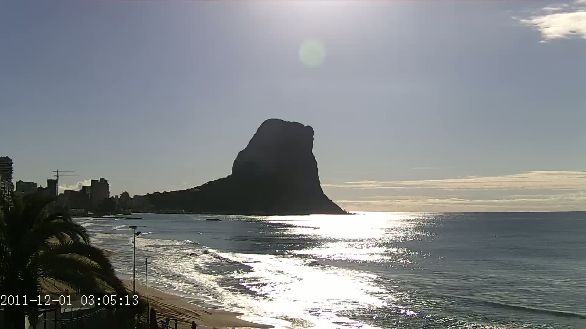 A large, dark, solitary mountain rises from the ocean on a sunny day with a partly cloudy sky, as waves lap a sandy shore lined with buildings and palm trees.
