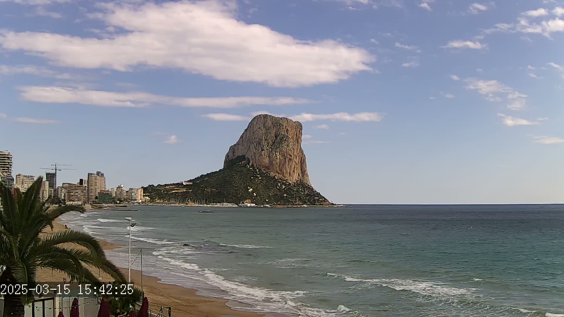 A large, rocky outcrop rises from the sea under a partly cloudy sky, with a sandy beach and buildings visible in the foreground.