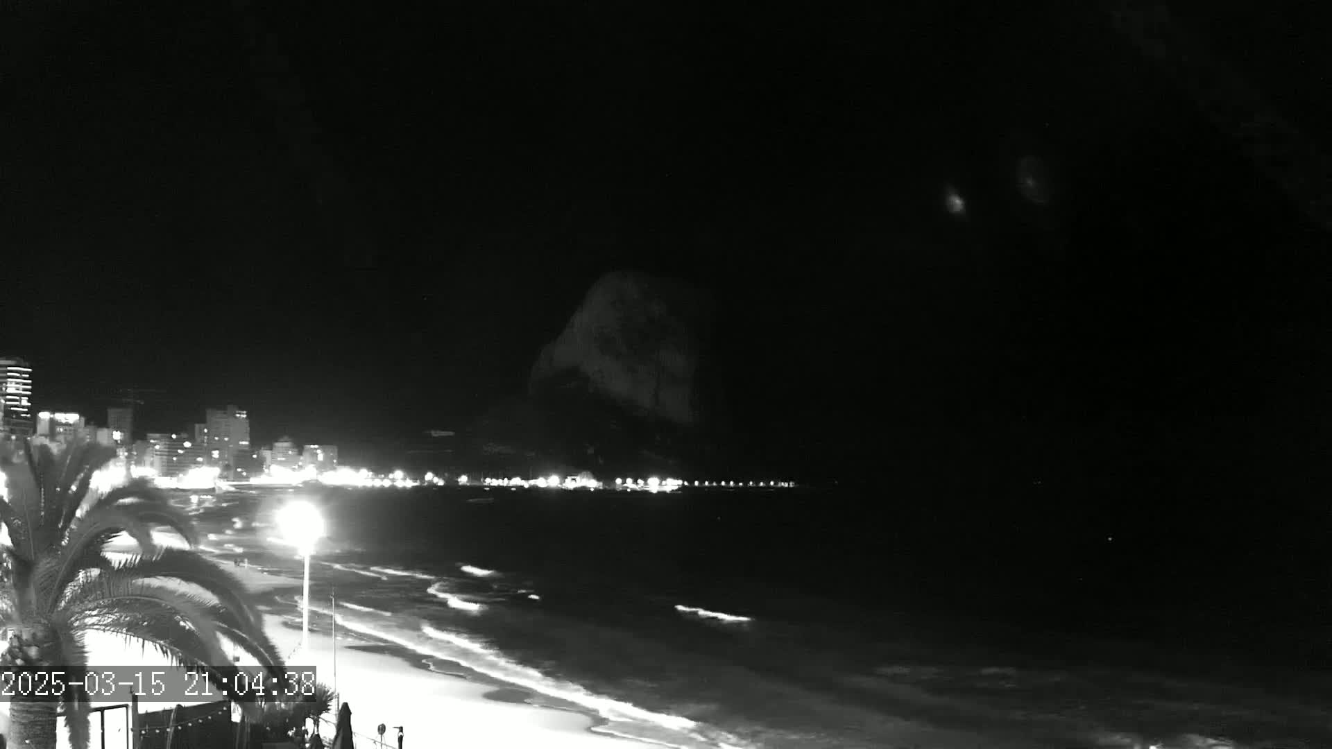 A nighttime beach scene shows gently lapping waves on a sandy shore, illuminated buildings and streetlights along the coast, and a dark mountain in the distance under a clear, dark sky.