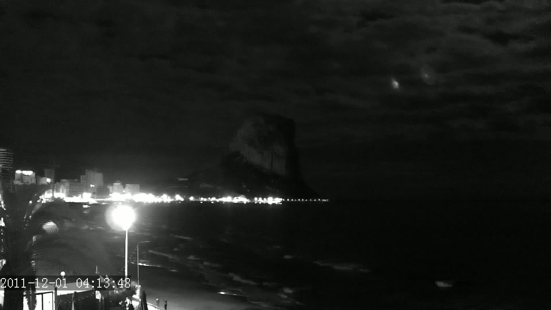 A nighttime, cloudy coastal scene shows a dark, rocky cliff in the distance, illuminated city lights along a beach, and gently waving water.