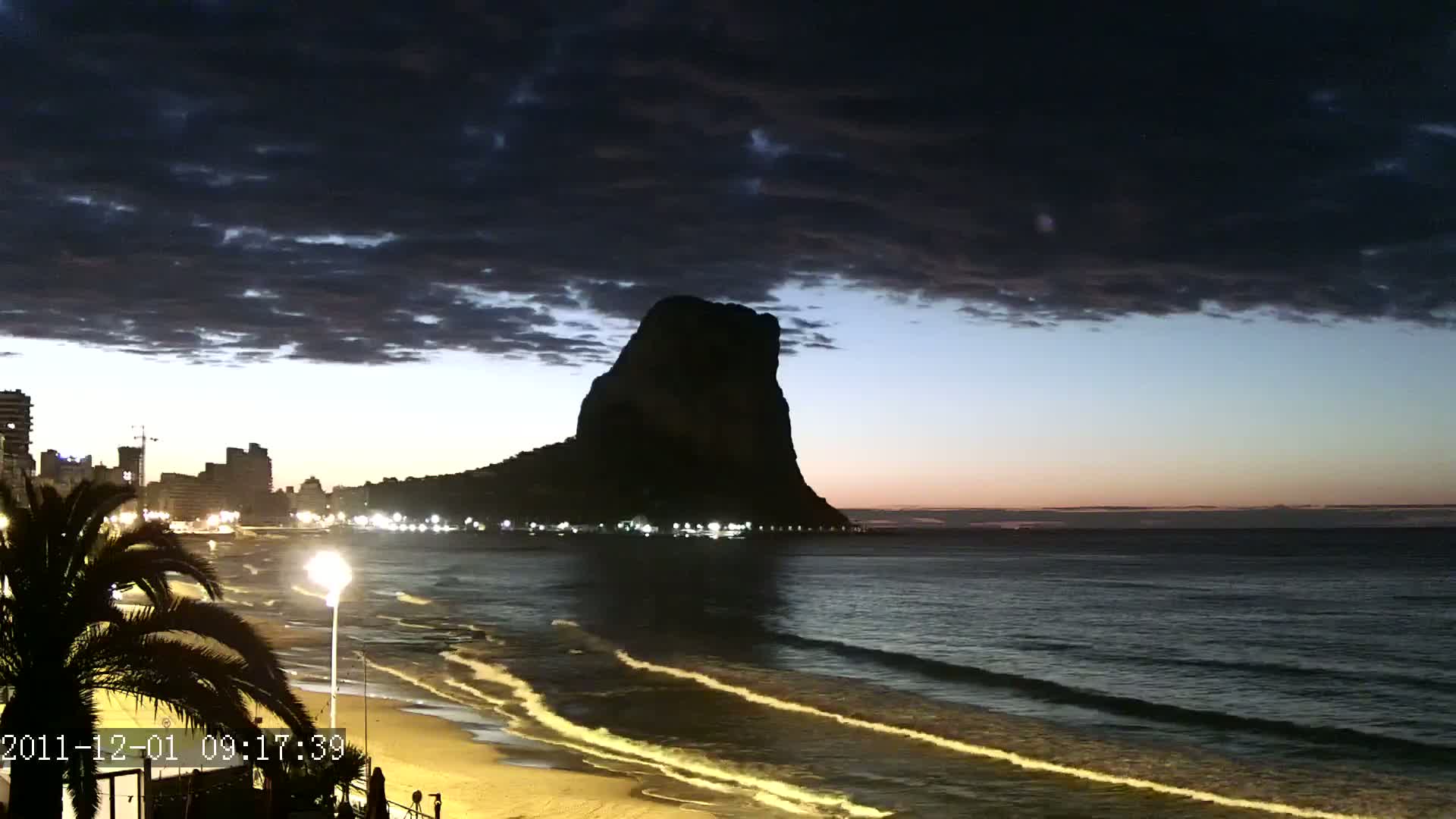 A dark, cloudy twilight sky overlooks a beach with gentle waves lapping the shore, a city skyline to the left, and a large, silhouetted rock formation in the distance.