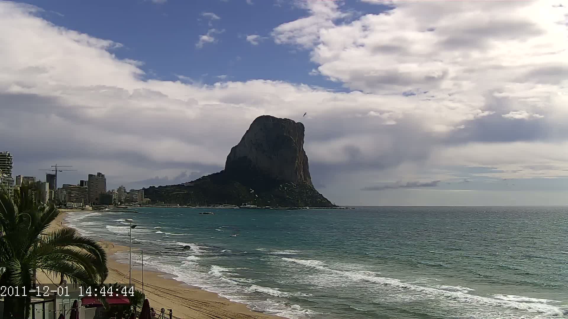 A large, dark rock formation rises from the sea under a partly cloudy sky, with a sandy beach and buildings visible in the foreground.