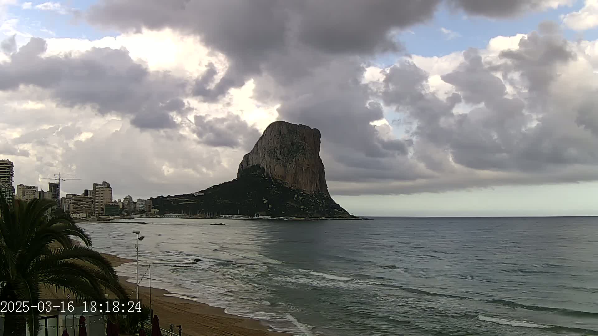 A large, dark rock formation rises from the sea under a cloudy sky, with a beach and buildings visible in the foreground.
