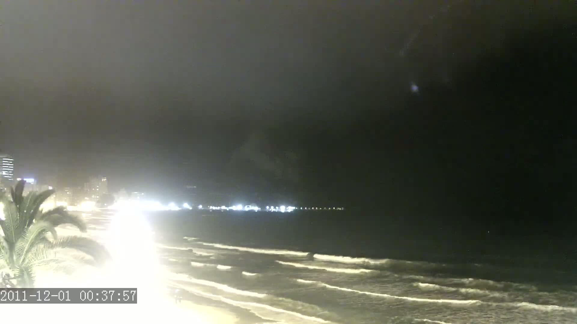 A nighttime view of a beach with gently rolling waves under a dark, overcast sky, showing city lights in the distance.