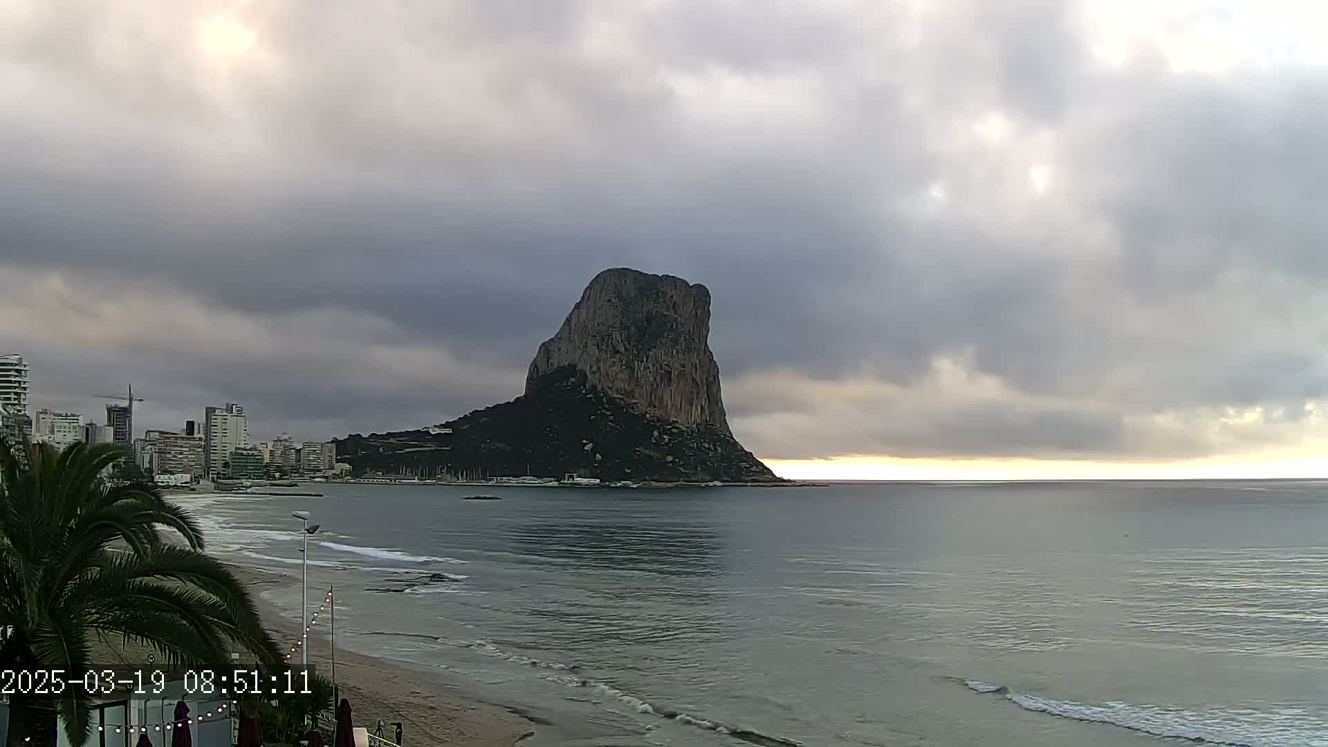 A large, dark rock formation rises from the sea under a cloudy sky, with a beach and cityscape visible in the foreground.