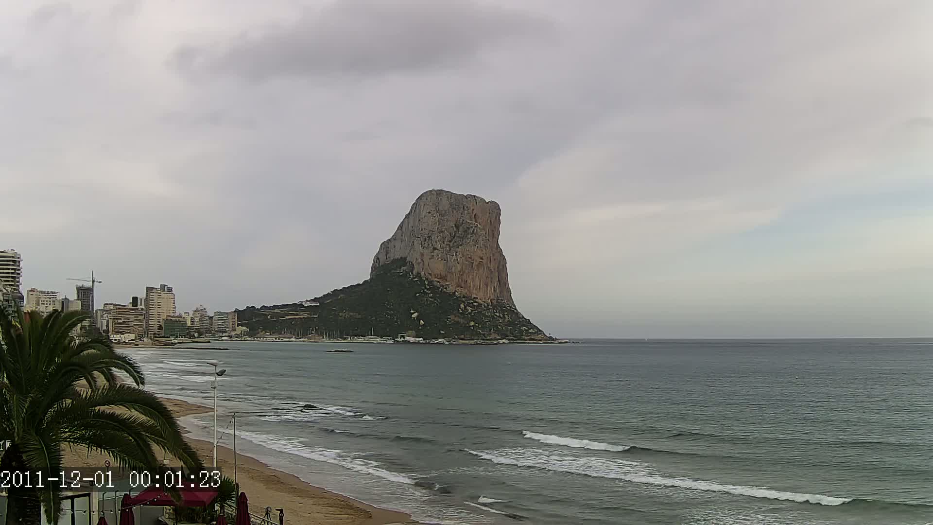 A large, rocky mountain rises from the sea under an overcast sky, with a sandy beach and buildings visible in the foreground.