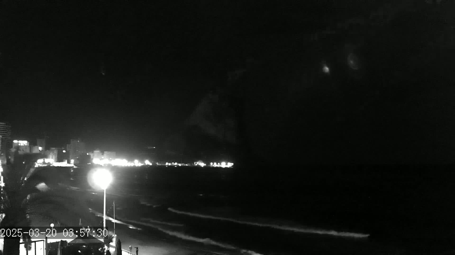 A nighttime beach scene shows a dark ocean with small waves lapping the shore, illuminated by lights from buildings in the distance under a mostly clear, dark sky.