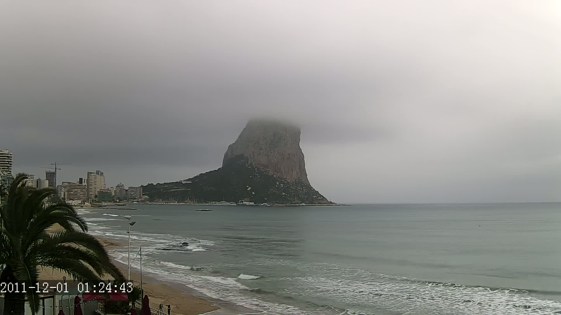 A large, fog-shrouded rock formation is visible across a calm, gray sea from a beach with buildings in the background under an overcast sky.