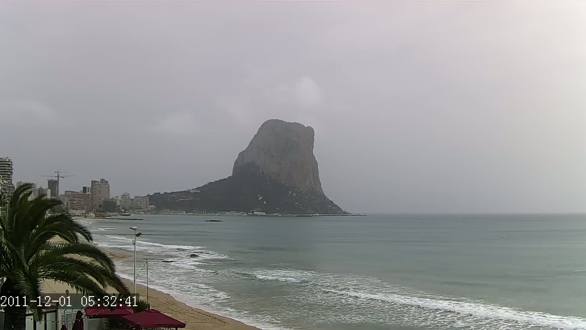 A large, dark rock formation rises from the sea on an overcast day, with a sandy beach and buildings visible in the foreground.