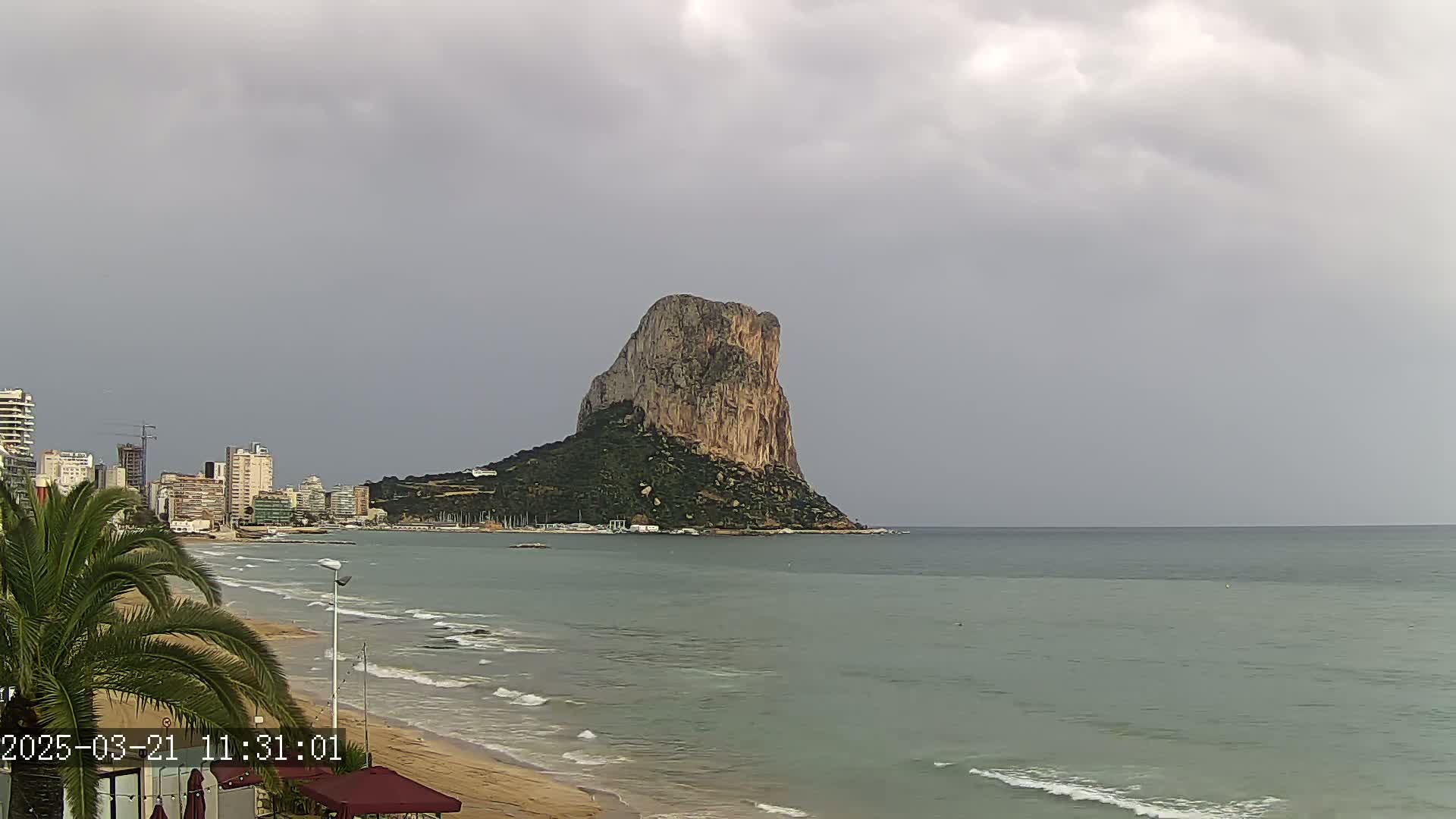 A large, rocky island rises from the calm, gray sea under an overcast sky, with a sandy beach and a line of buildings visible in the foreground.