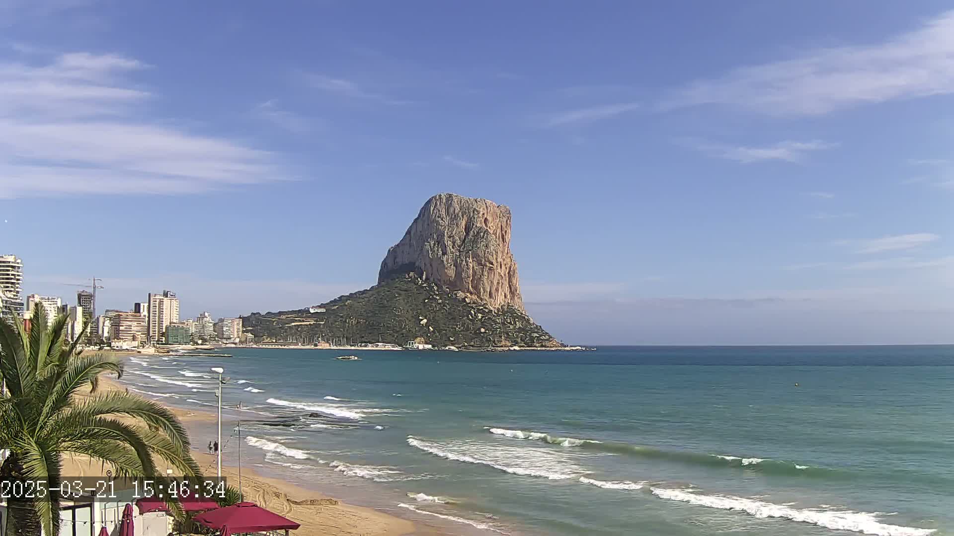 A sandy beach with gentle waves laps the shore, overlooking a city skyline and a large, rocky, isolated island under a mostly clear blue sky.