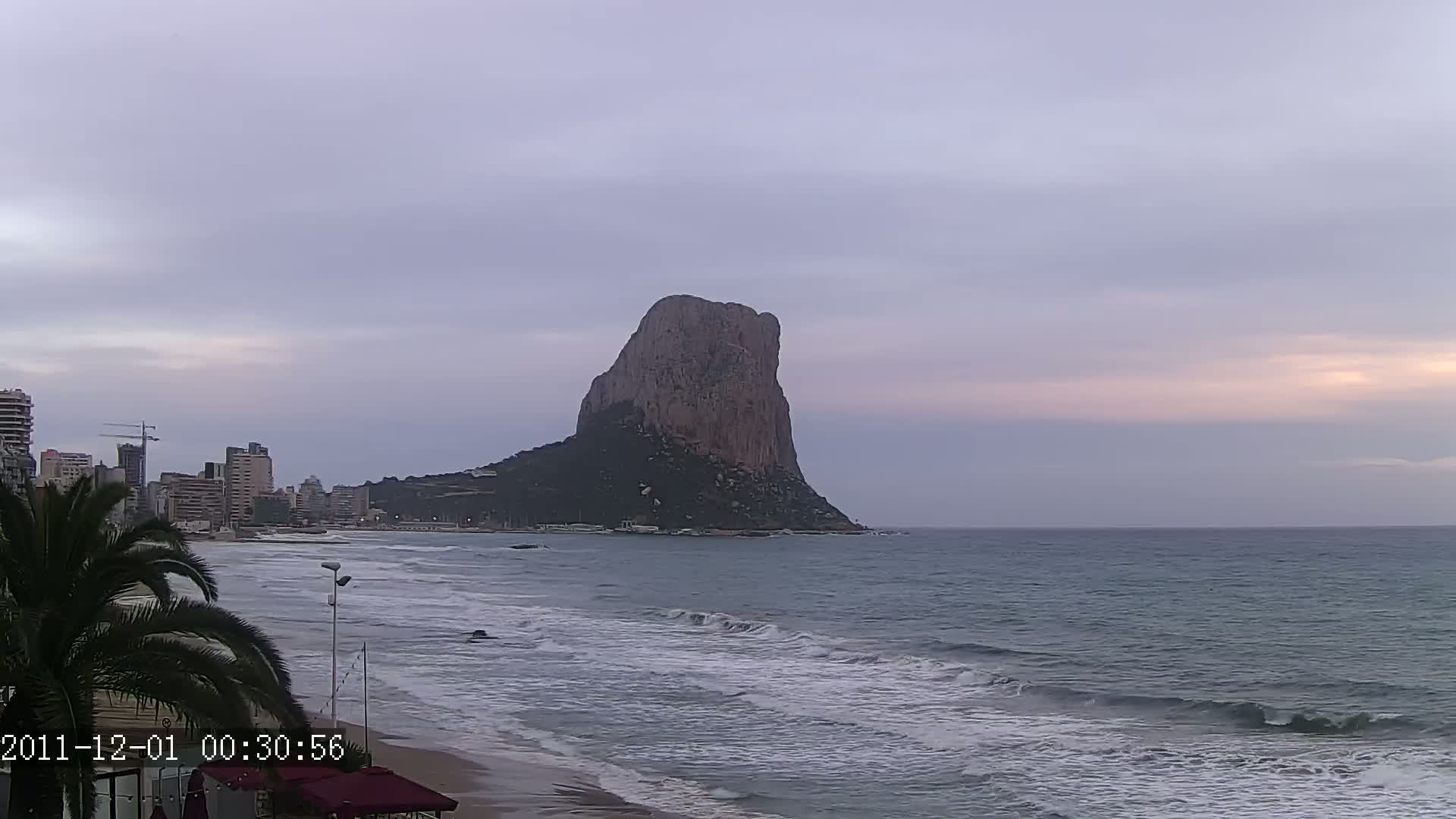 A large, dark rock formation rises from the ocean under a cloudy, pale sky, with a city and beach visible in the foreground.
