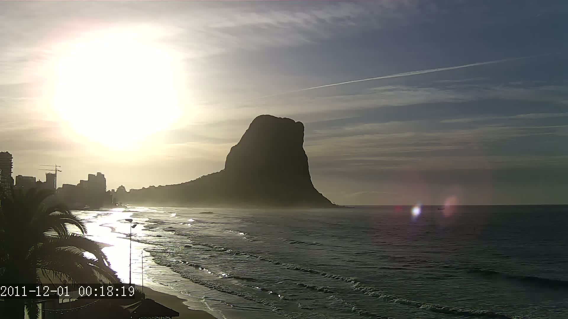 A large, dark mountain rises behind a beach with gentle waves under a bright sun in a mostly clear sky.