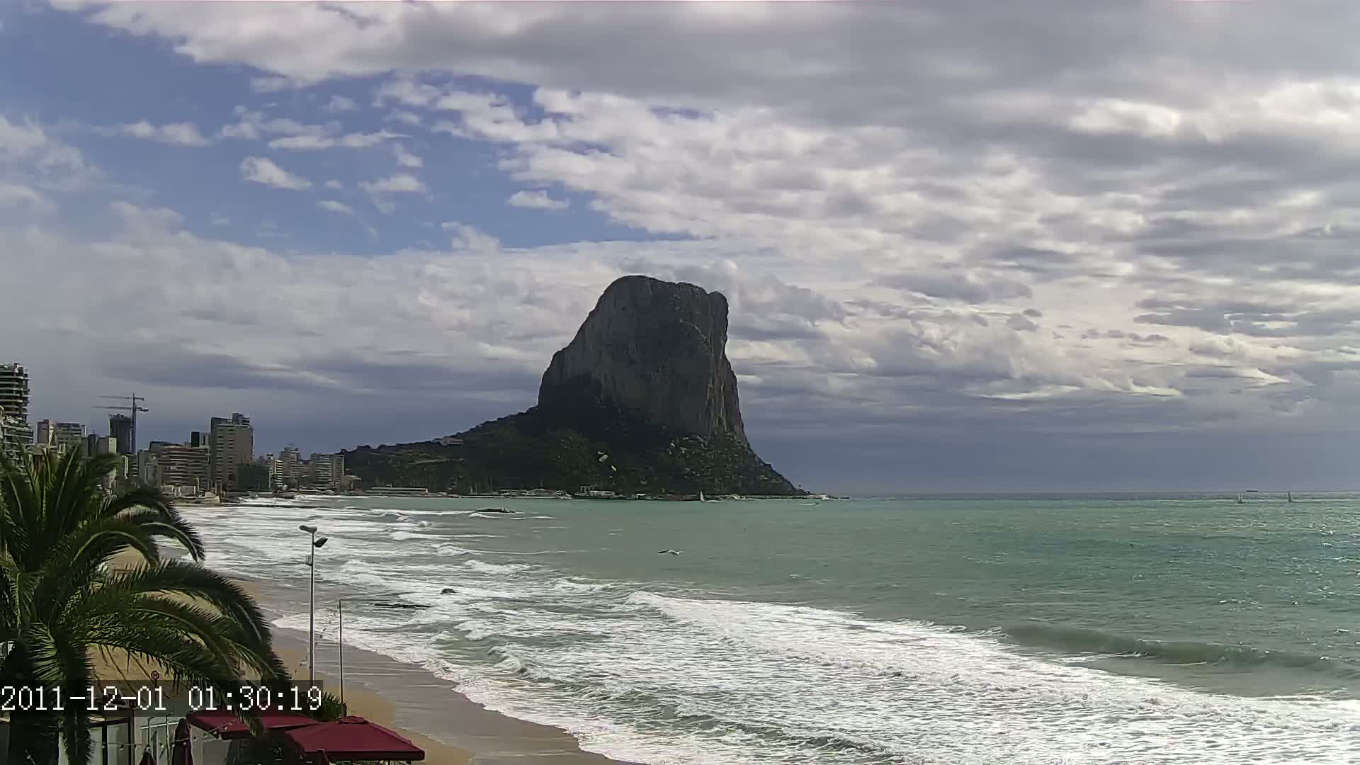 A large, dark rock formation rises from the sea under a partly cloudy sky, with waves breaking on a sandy beach in the foreground and buildings visible in the distance.