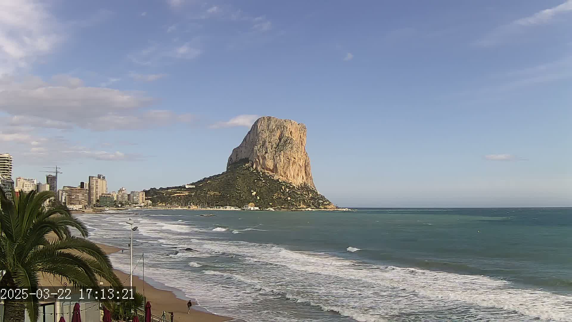 A large, light-brown rock formation rises from the sea near a sandy beach with gentle waves under a mostly clear sky.