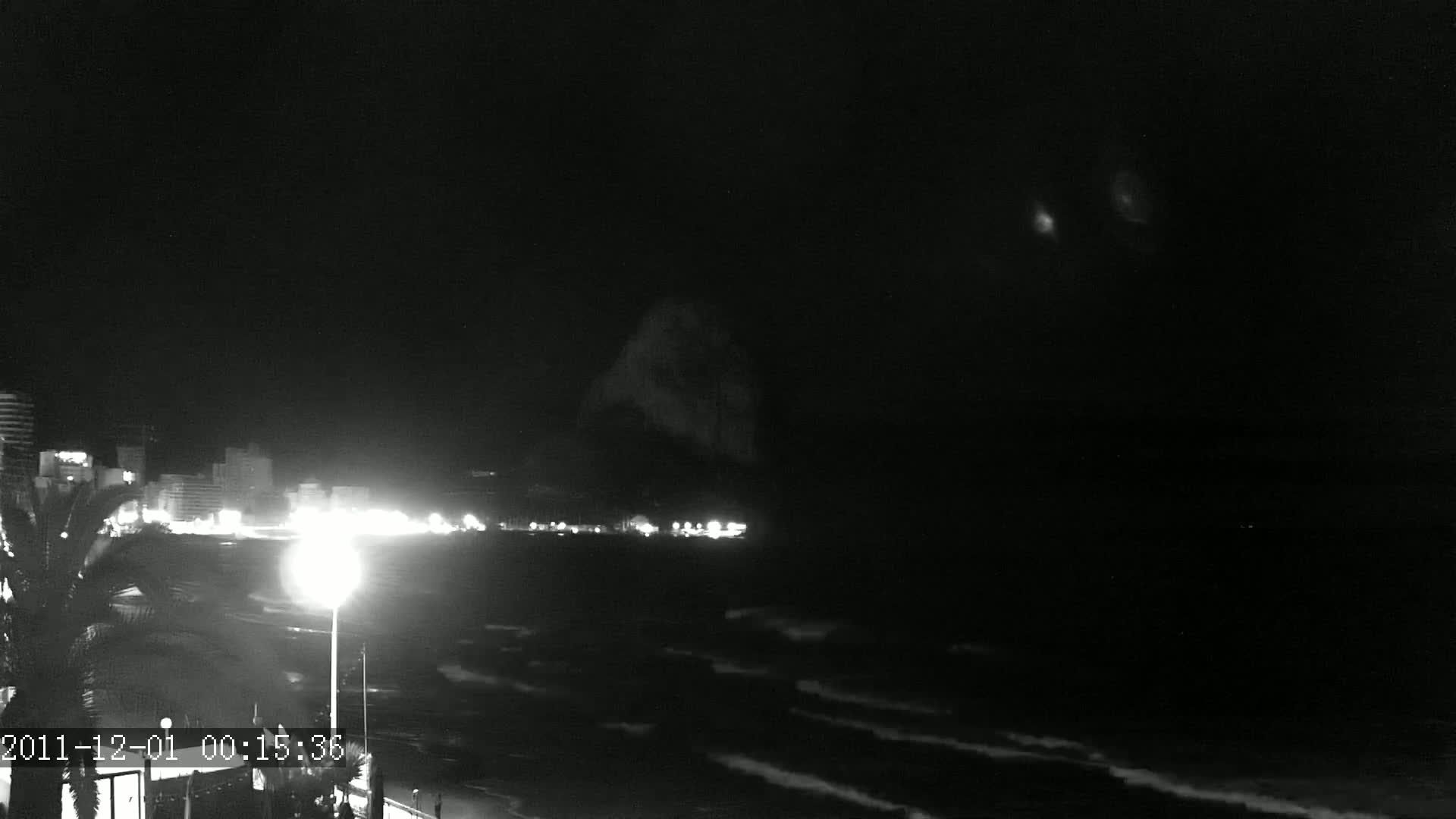 A nighttime, low-light beach scene shows a dark, wavy ocean, a line of illuminated buildings, and a large, dark landmass in the distance under a mostly clear sky.