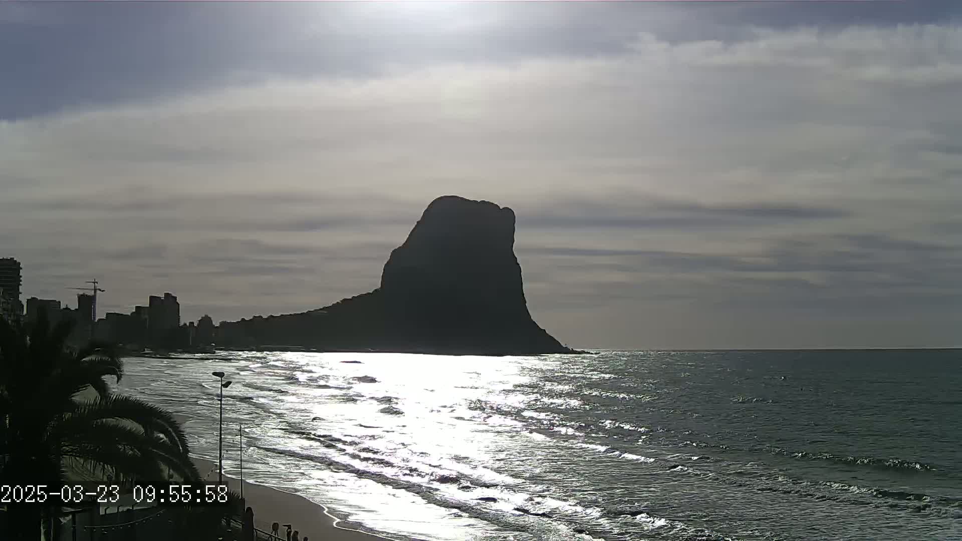 A large, dark mountain rises from the sea under a partly cloudy sky, with a beach and city skyline visible in the foreground.