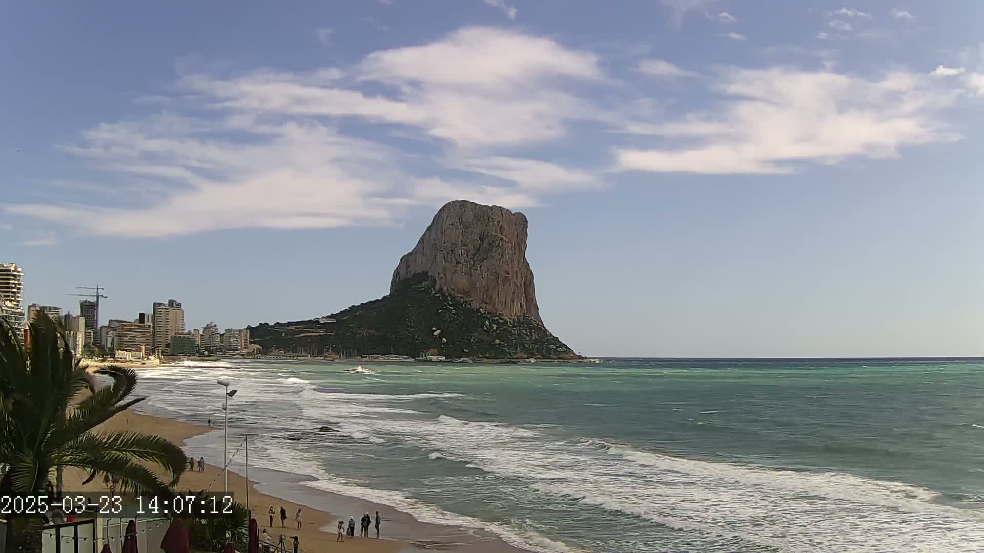 A sandy beach with a few people on it is next to a wavy ocean, with a large, rocky, cliff-like landmass in the distance under a partly cloudy sky.