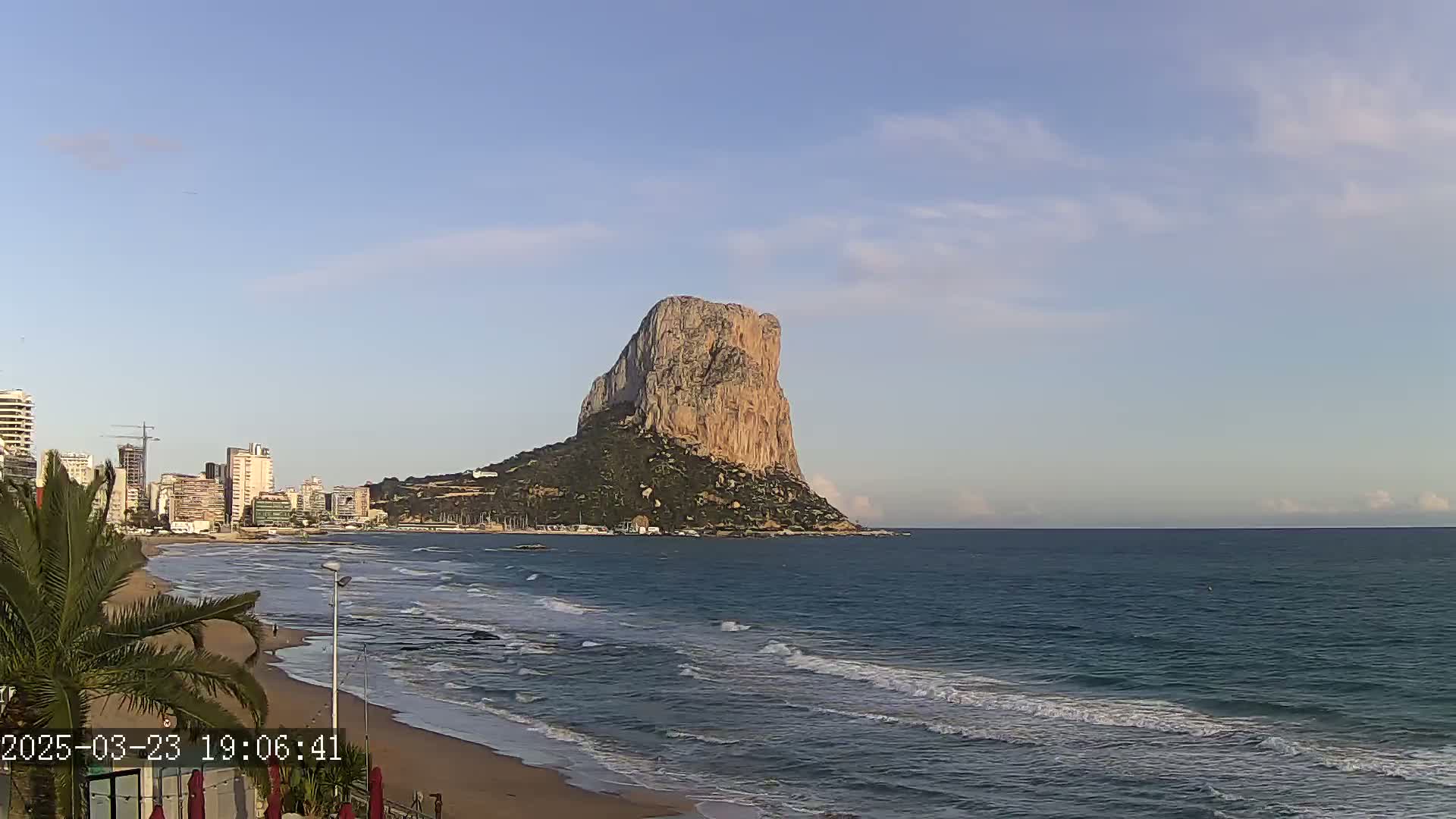 A large, tan rock formation rises from the sea under a partly cloudy sky, with a sandy beach and cityscape visible in the foreground.