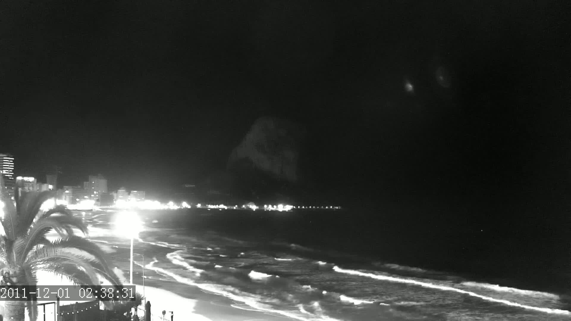 A nighttime beach scene shows gently rolling waves lapping on the shore, with city lights illuminating the coastline and a dark mountain in the background under a clear, dark sky.