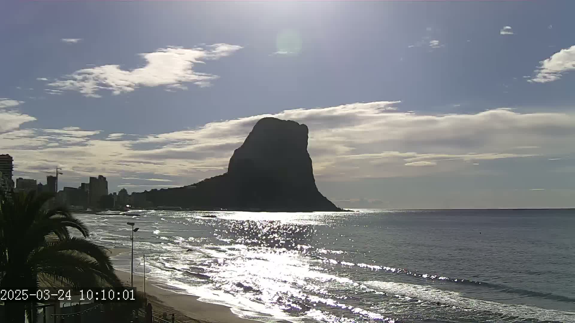 A large, dark mountain rises from the ocean under a partly cloudy sky, with a sandy beach in the foreground and a city skyline visible in the distance.