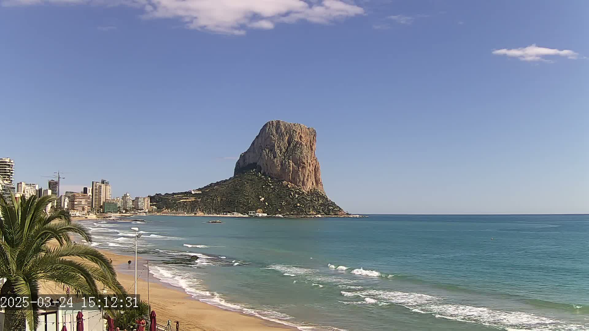 A sandy beach with gentle waves laps the shore, under a mostly clear blue sky, with a large, rocky island visible in the distance and buildings lining the coast.