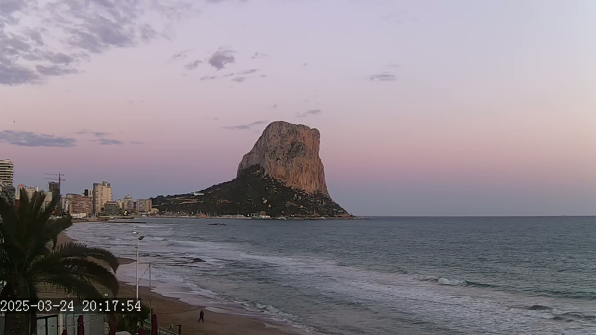 A large, rocky island rises from the sea under a pale pink and purple twilight sky, with a sandy beach and city skyline visible in the foreground.
