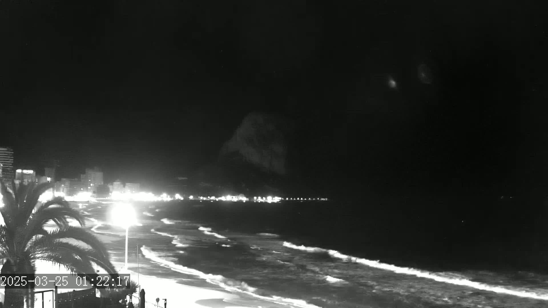 A nighttime, black-and-white view of a beach with gently rolling waves, a shoreline with illuminated buildings, and a dark, possibly cloudy, sky.