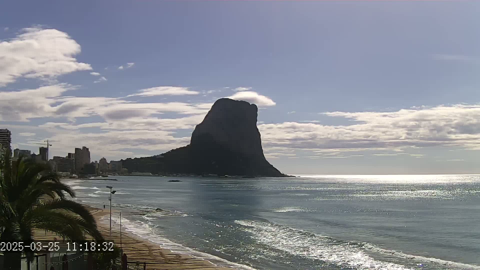 A large, dark mountain rises from the sea under a partly cloudy sky, with a sandy beach and buildings visible in the foreground.
