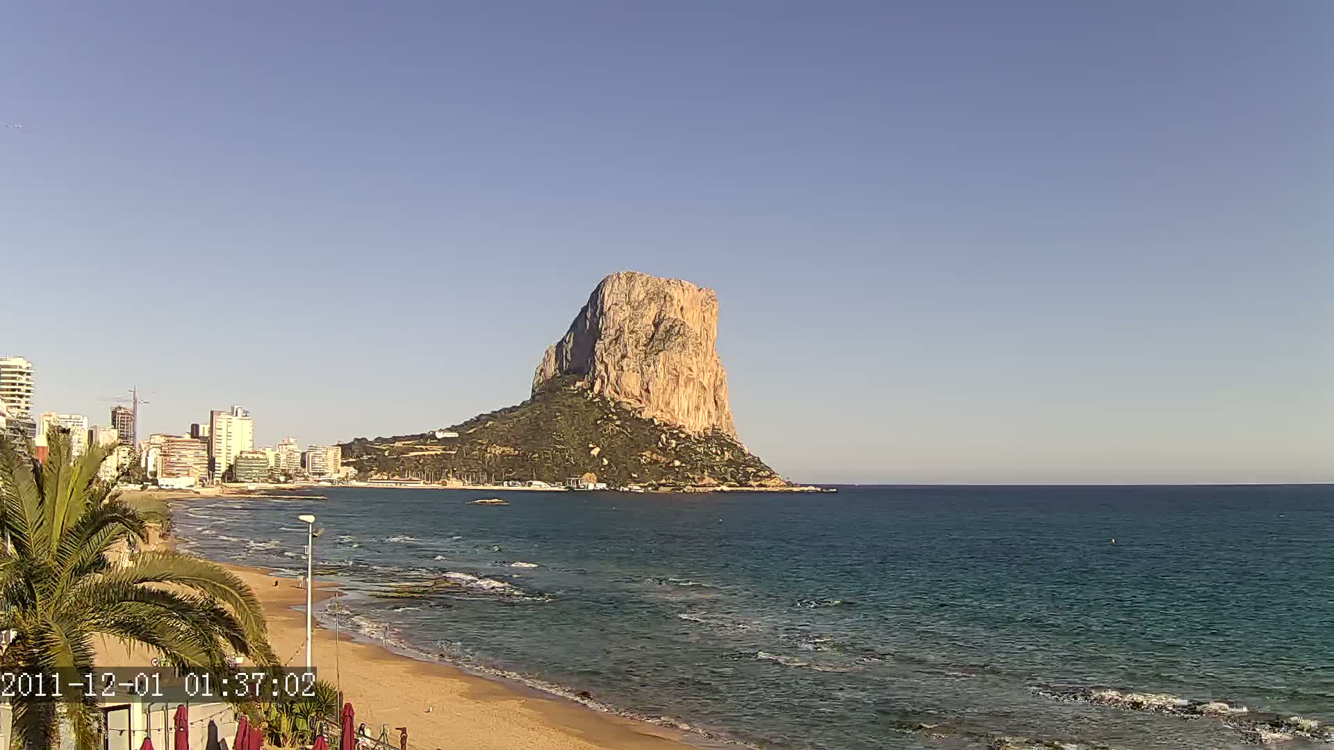 A large, tan rock formation rises from the sea near a sandy beach and a line of buildings under a clear, bright sky.