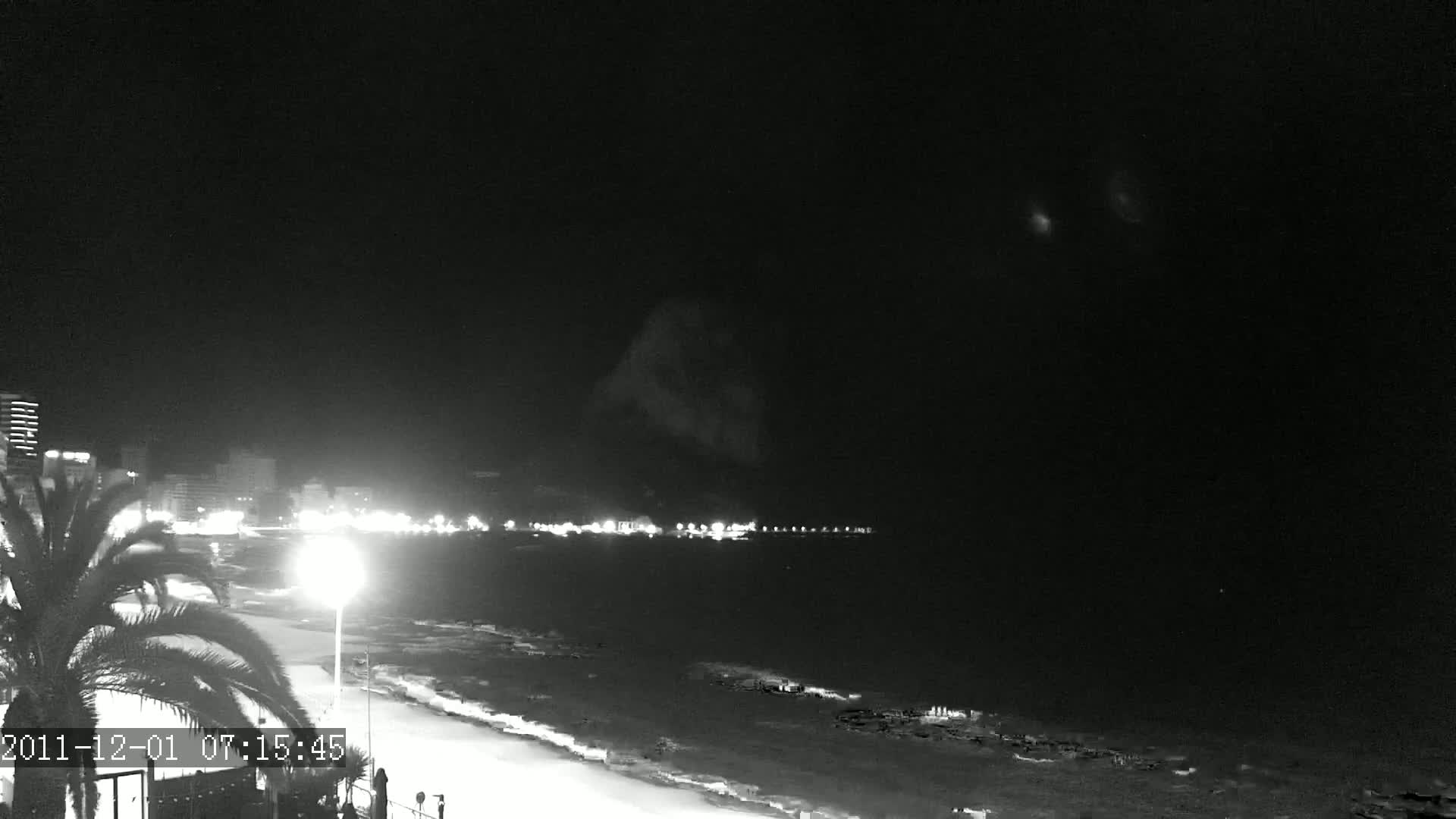 A nighttime, black and white view of a beach at night with a cityscape in the distance under a cloudy sky.