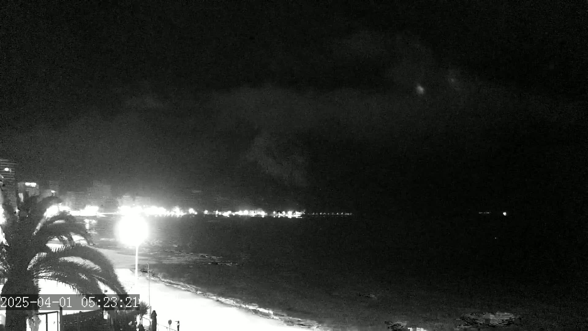 A nighttime view of a beach with a calm sea, a dimly lit boardwalk, and a distant city skyline under a cloudy sky.