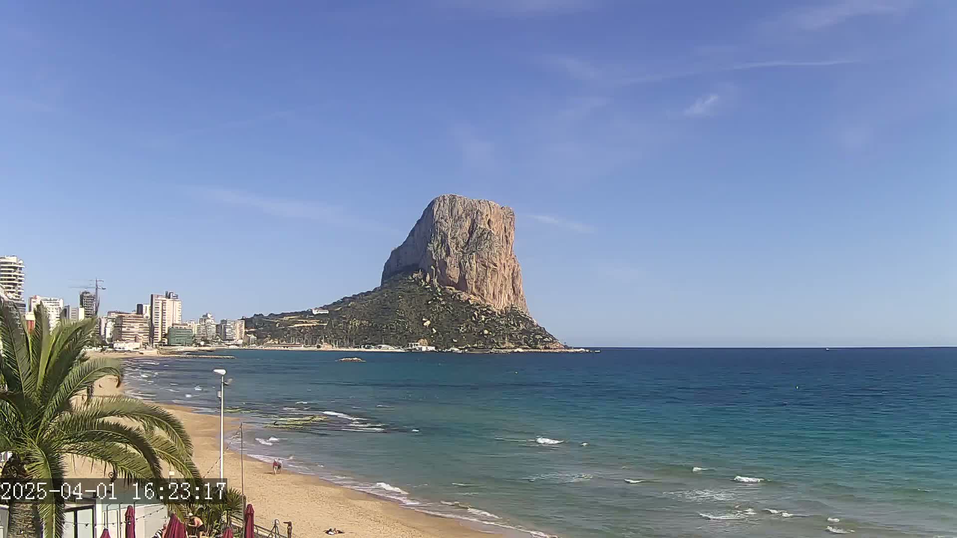 A sandy beach with calm, blue water stretches towards a large, imposing rock formation under a clear blue sky.