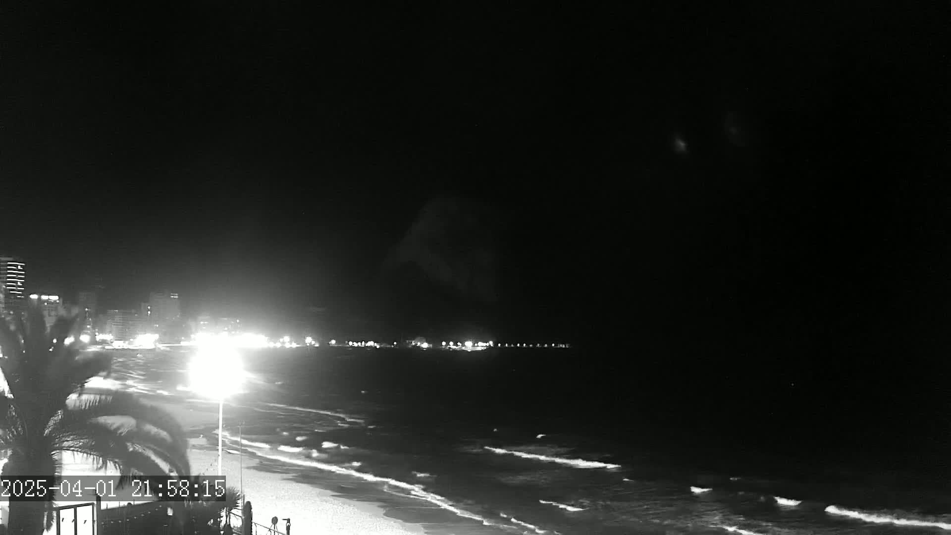 A nighttime view of a beach with gently lapping waves, illuminated by lights from buildings in the distance under a clear, dark sky.