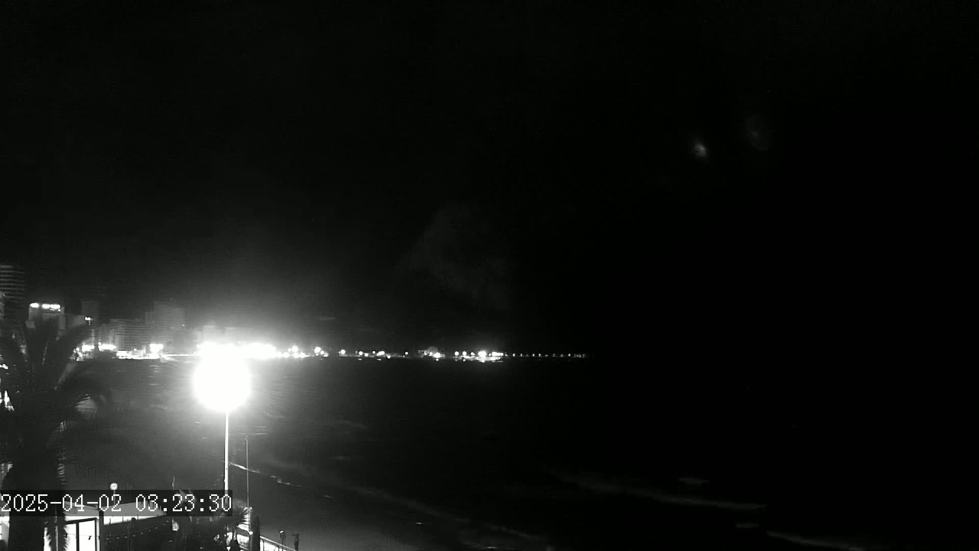A nighttime, low-light view of a dark ocean shore with distant city lights and a bright lamp in the foreground.