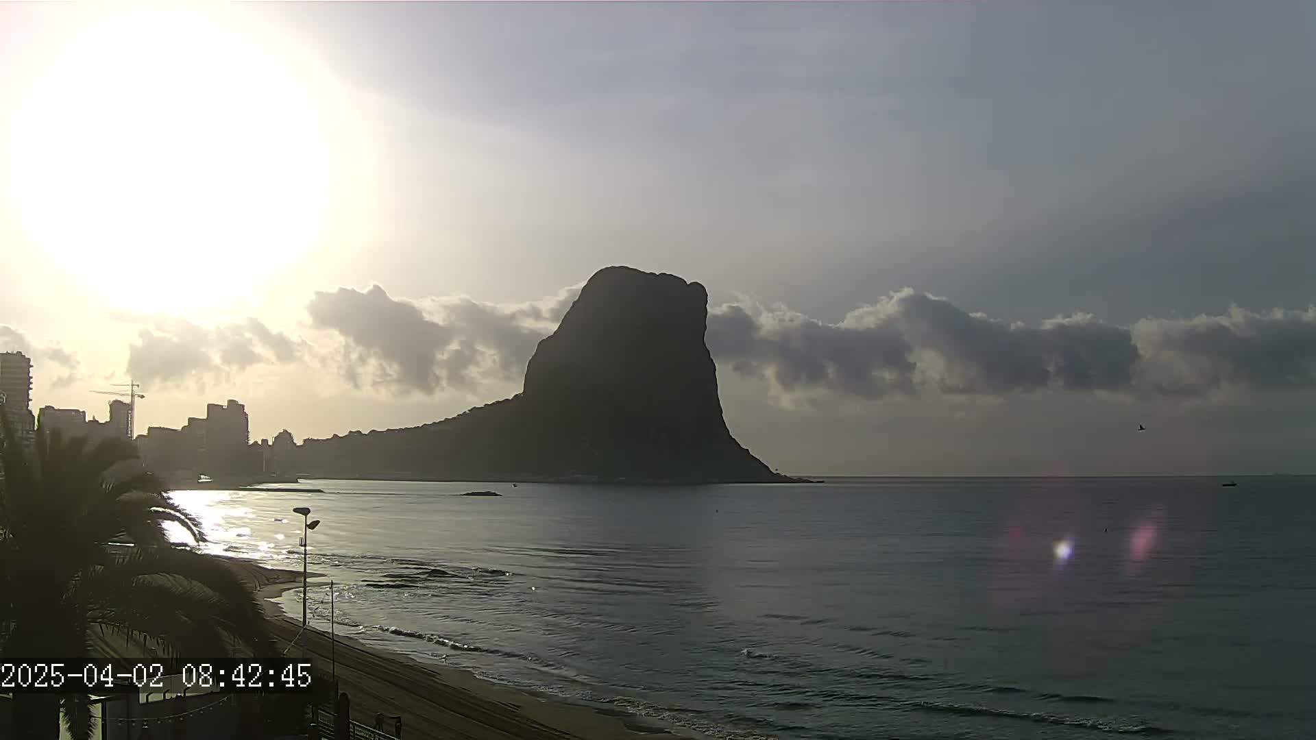 A large, dark rock formation rises from the sea under a partly cloudy sunrise, with a beach and city skyline visible in the foreground.