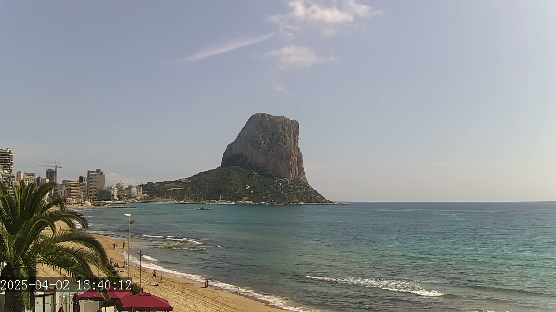 A large, rocky island rises from a calm, teal sea, a sandy beach with a few people stretches in the foreground, and a city skyline is visible in the distance under a mostly clear sky.