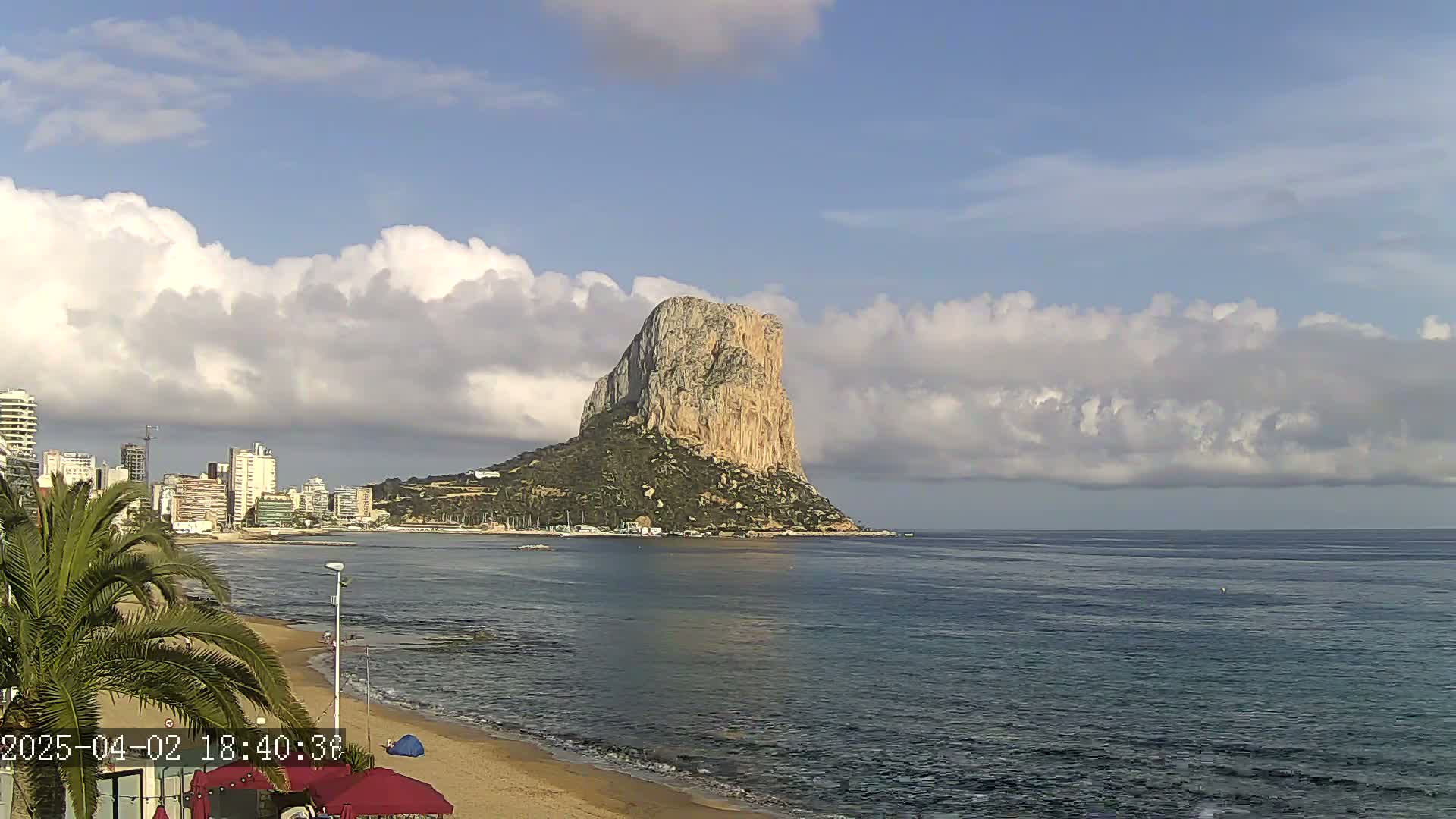A large, rocky island rises from the sea under a partly cloudy sky, with a sandy beach and buildings visible in the foreground.
